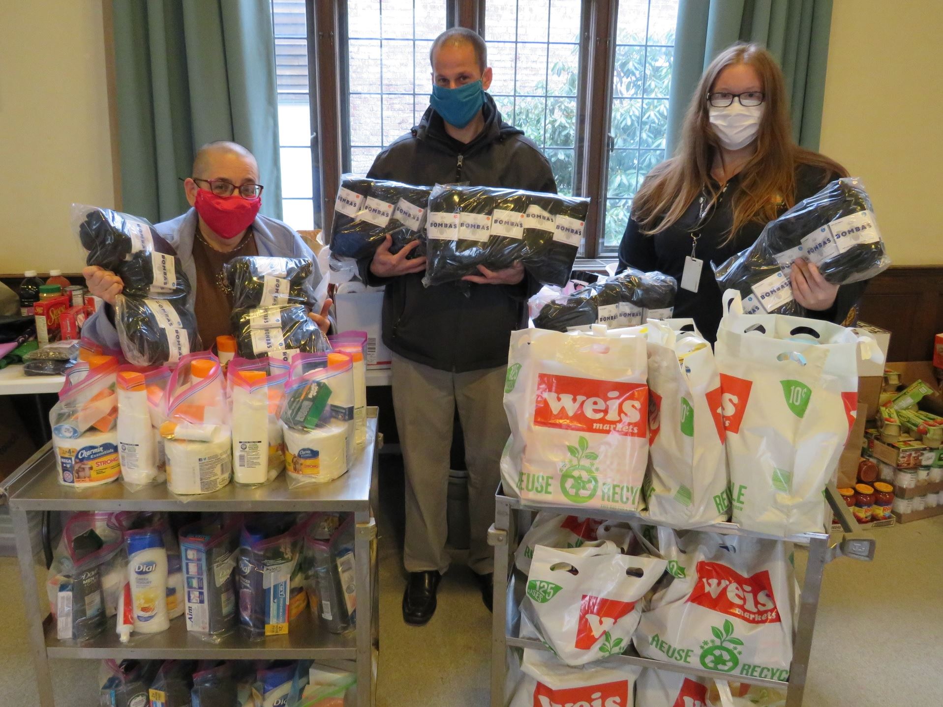 Three people holding donated Bombas Socks, surrounded by pantryand sundry items for distribution.